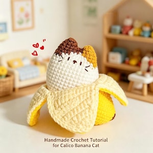 May include: A handmade crochet calico cat plush emerging from a yellow banana peel. The cat has white, brown, and orange yarn details with embroidered facial features. Red heart icons float above. Text reads: Handmade Crochet Tutorial for Calico Banana Cat.