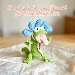 May include: A green crocheted lizard toy with a light blue frill, large white eyes with black pupils, and a long pink tongue. The text above the figure reads: Handmade Crochet Tutorial for Crazy Lizard Tom. The lizard has four limbs.