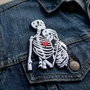 May include: An embroidered patch featuring two white skeletons embracing, with one skeleton positioned behind the other. The skeletons are rendered in white thread against a black background, and a single bright red heart is centered on the chest.