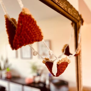 May include: A handmade crochet garland featuring triangular slices of pie. The pie slices are a deep red-brown with a tan crust, strung together with white yarn. The garland is hanging in front of a decorative mirror.