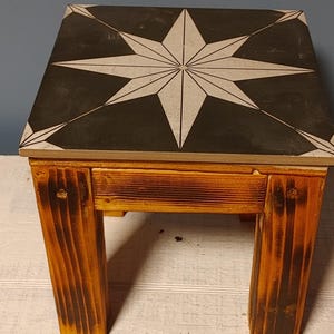May include: A small wooden side table with a square top. The tabletop is black with a white compass rose design. The legs and frame are made of stained wood. The table is approximately 12 inches tall.