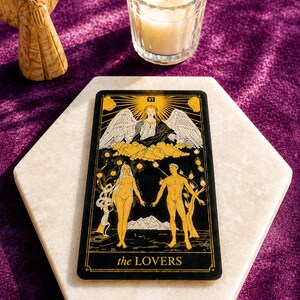 May include: A tarot card with the text "the LOVERS" on a white marble surface. The card features illustrations of a man and woman, with an angel above them. A lit candle in a glass holder and a small angel statue are also in the image.