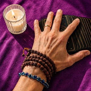 May include: A lit candle in a clear glass holder, a deck of tarot cards, and two bracelets on a purple surface. One bracelet is made of brown beads, and the other has blue stones. A hand with a gold ring rests on the cards.