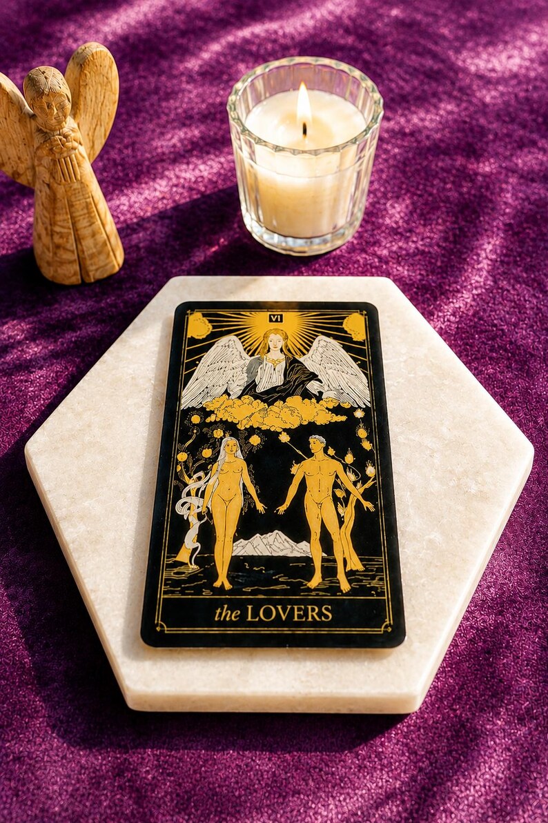 May include: A tarot card with the text "the LOVERS" on a white marble surface, with a lit candle and a small angel statue in the background. The card features illustrations of a man and woman, with an angel above them.
