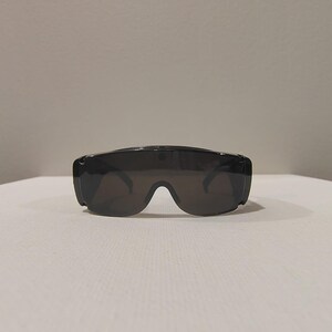 Puede incluir: Gafas de sol negras con un dise&ntilde;o envolvente ancho. Las lentes son oscuras y la montura es gruesa y s&oacute;lida. Las gafas de sol est&aacute;n sobre una superficie blanca, con un fondo liso.