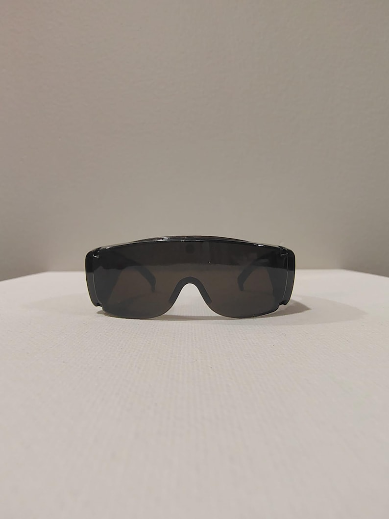 Puede incluir: Gafas de sol negras con un dise&ntilde;o envolvente ancho. Las lentes son oscuras y la montura es gruesa y s&oacute;lida. Las gafas de sol est&aacute;n sobre una superficie blanca, con un fondo liso.