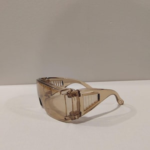 Puede incluir: Gafas de seguridad transparentes de color marr&oacute;n claro con un dise&ntilde;o envolvente. Las gafas tienen una montura curvada y protectores laterales con ranuras de ventilaci&oacute;n. Las lentes est&aacute;n tintadas y las patillas se curvan sobre las orejas.