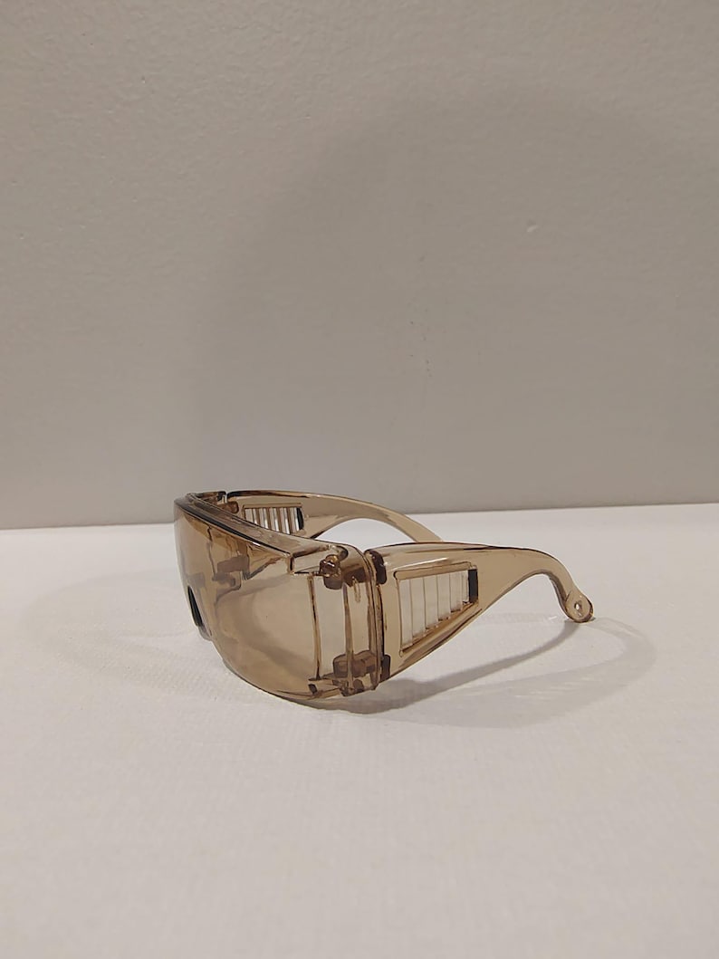 Puede incluir: Gafas de seguridad transparentes de color marr&oacute;n claro con un dise&ntilde;o envolvente. Las gafas tienen una montura curvada y protectores laterales con ranuras de ventilaci&oacute;n. Las lentes est&aacute;n tintadas y las patillas se curvan sobre las orejas.