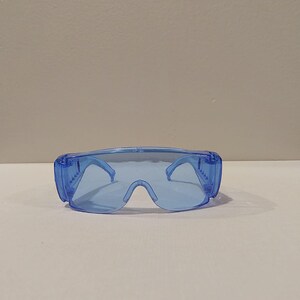 Puede incluir: Un par de gafas de seguridad azules transparentes. Las gafas tienen un dise&ntilde;o envolvente con una montura gruesa y lentes transparentes. Las patillas son finas y se curvan alrededor de las orejas. Las gafas est&aacute;n dise&ntilde;adas para proteger los ojos.