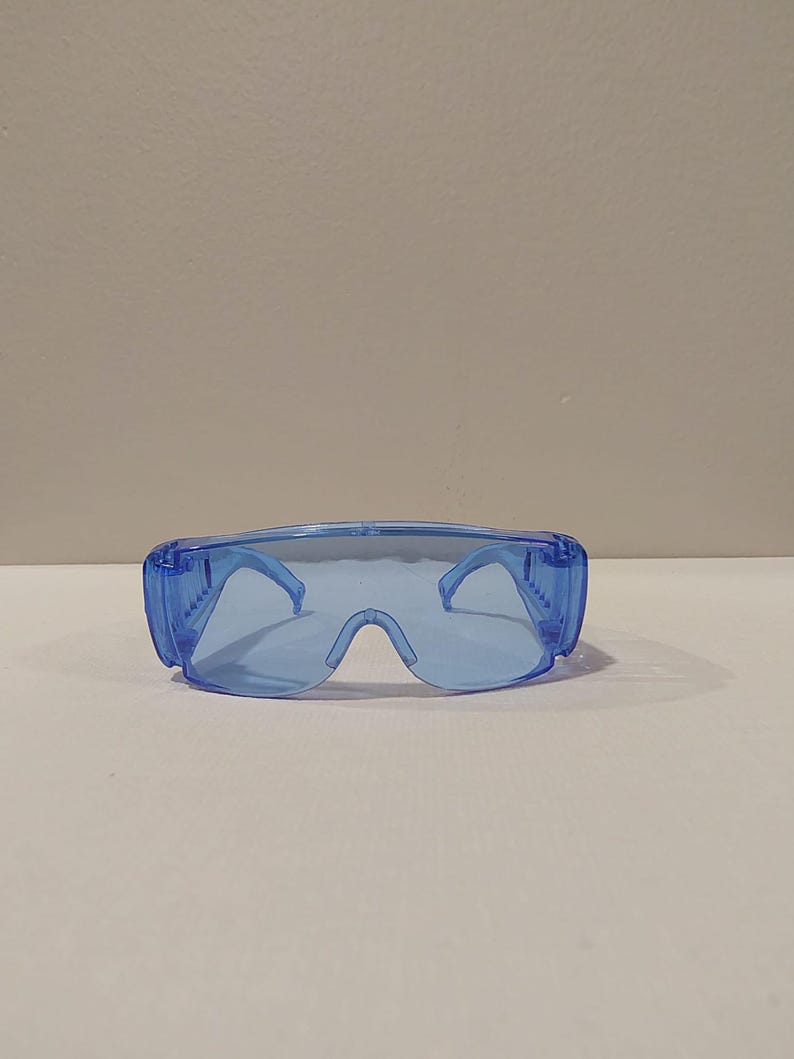 Puede incluir: Un par de gafas de seguridad azules transparentes. Las gafas tienen un dise&ntilde;o envolvente con una montura gruesa y lentes transparentes. Las patillas son finas y se curvan alrededor de las orejas. Las gafas est&aacute;n dise&ntilde;adas para proteger los ojos.