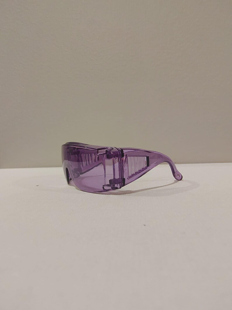 Puede incluir: Un par de gafas de seguridad moradas transparentes. Las gafas tienen un dise&ntilde;o envolvente con una patilla curva para un ajuste seguro. Las lentes y la montura son de color morado s&oacute;lido y transl&uacute;cido, ofreciendo protecci&oacute;n ocular.