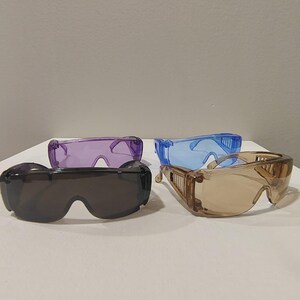 Puede incluir: Cuatro pares de gafas de seguridad en varios colores: gris oscuro, morado, azul y marr&oacute;n claro. Las gafas tienen lentes transparentes y protectores laterales. Las gafas est&aacute;n dispuestas sobre una superficie blanca.
