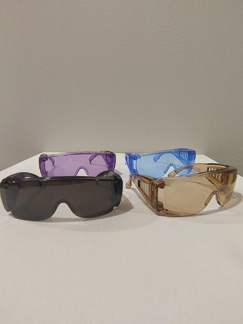 Puede incluir: Cuatro pares de gafas de seguridad en varios colores: gris oscuro, morado, azul y marr&oacute;n claro. Las gafas tienen lentes transparentes y protectores laterales. Las gafas est&aacute;n dispuestas sobre una superficie blanca.