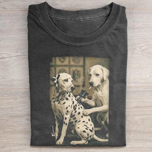 May include: Dark gray t-shirt featuring a sepia-toned image of two dogs. One is a Dalmatian getting a tattoo from a yellow Labrador. The Labrador is holding a tattoo gun and a cigarette. The Dalmatian has black spots.