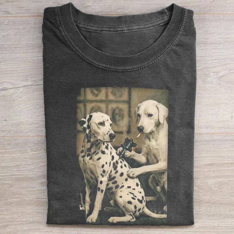 May include: Dark gray t-shirt featuring a sepia-toned image of two dogs. One is a Dalmatian getting a tattoo from a yellow Labrador. The Labrador is holding a tattoo gun and a cigarette. The Dalmatian has black spots.