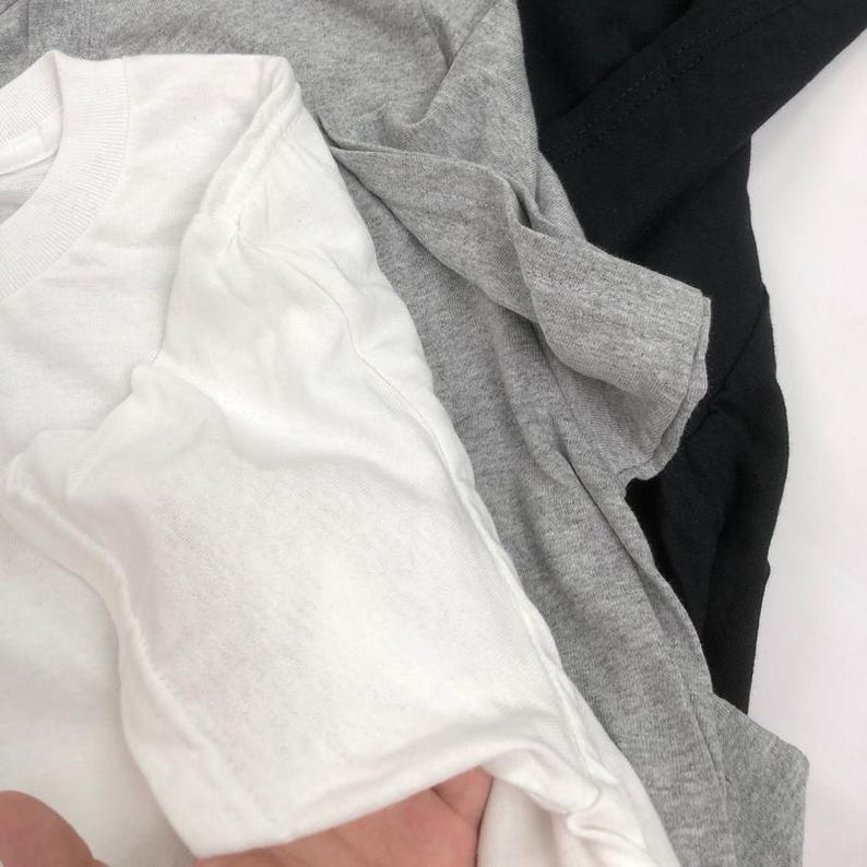 May include: A close-up shot of three folded t-shirts in white, gray, and black. The white t-shirt is in the foreground, with the gray and black shirts layered behind it. The fabric appears soft and slightly wrinkled.