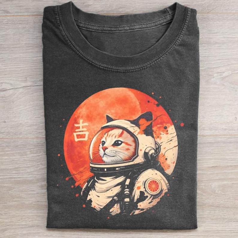 May include: Dark gray t-shirt featuring a cat in an astronaut helmet against a red planet backdrop. The design includes Japanese characters and a retro space theme. The cat is orange and white.