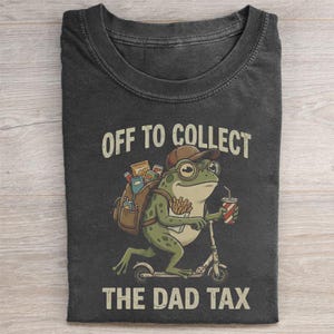 May include: Dark gray t-shirt featuring a cartoon frog riding a scooter. The frog wears glasses and a cap, carrying a backpack and a drink. Text reads "OFF TO COLLECT THE DAD TAX."