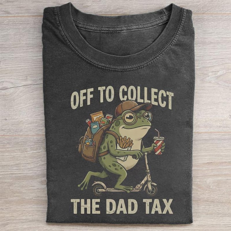 May include: Dark gray t-shirt featuring a cartoon frog riding a scooter. The frog wears glasses and a cap, carrying a backpack and a drink. Text reads "OFF TO COLLECT THE DAD TAX."