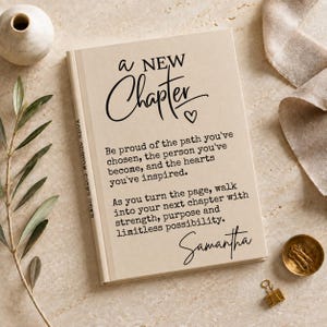 May include: A beige hardcover book with black text that reads: A NEW Chapter. Be proud of the path you've chosen, the person you've become, and the hearts you've inspired. As you turn the page, walk into your next chapter with strength, purpose and limitless possibility. Samantha.