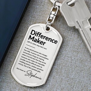 May include: A silver-toned metal dog tag keychain featuring the black printed text Difference Maker, a phonetic pronunciation, a definition of the term, and the name Stephanie at the bottom, attached to a silver metal key ring with a metal key.