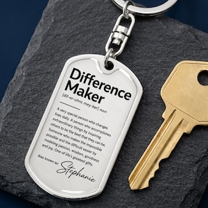 May include: A silver-toned metal dog tag keychain featuring the black printed text Difference Maker, a phonetic pronunciation, a definition of the term, and the name Stephanie at the bottom, attached to a circular split ring and a gold-toned key.