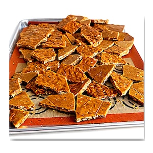 May include: A tray of peanut brittle, broken into irregular pieces. The brittle is a golden brown color, with visible peanuts throughout. The pieces are arranged on a red silicone baking mat.
