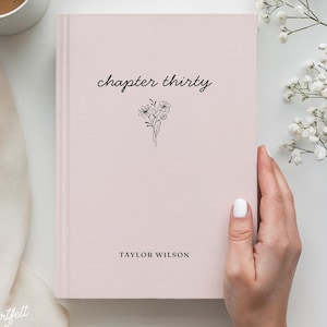 Puede incluir: Un libro rosa claro con las palabras "chapter thirty" en cursiva y una pequeña ilustración floral. El nombre del autor, "TAYLOR WILSON", está impreso debajo. Una mano sostiene el libro, con una taza de café y flores en el fondo.