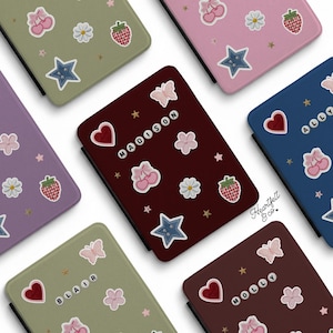 May include: A collection of rectangular e-reader cases in various colors including olive green, pink, purple, dark red, and blue. Each case features embroidered patches of cherries, daisies, strawberries, stars, butterflies, hearts, and personalized names.