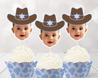 May include: Three cupcakes with white frosting and blue cow-print liners. Each cupcake is topped with a baby's face and a brown cowboy hat with a blue star and trim. The image is set against a blurred background.