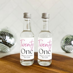 May include: Two small clear glass bottles with silver caps and white labels. The labels have pink script that says "Twenty One" and black text that says "Megyn's 21st Bday 06.12.XX".