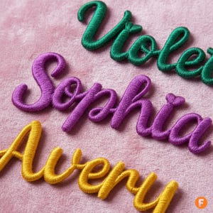 May include: Close-up of embroidered names in cursive script. The names are in various colors: green, purple, and yellow. The background is a soft pink fabric. The names are Violet, Sophia, and Avery.