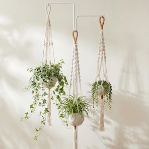 May include: Three macrame plant hangers with white ceramic pots and trailing greenery. The hangers are suspended from a white metal frame. The macrame features wooden beads and a tassel at the bottom, creating a bohemian aesthetic.
