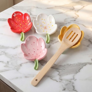 May include: A collection of four ceramic spoon rests in the shape of flowers. One is red with the name "Bella", one is white, one is pink, and one is yellow. A wooden spoon rests in the yellow spoon rest. The spoon rests have green stems.