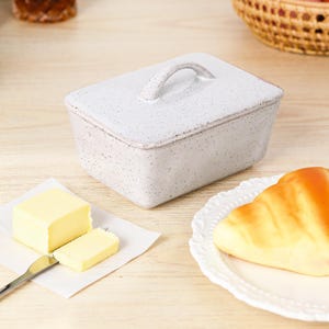 Könnte beinhalten: Eine gesprenkelte weiße Keramik-Butterdose mit einem gebogenen Griff auf dem Deckel. Ein Butterstück und eine Scheibe liegen auf einem kleinen Papierquadrat mit einem Buttermesser. Ein Croissant liegt auf einem weißen Teller.