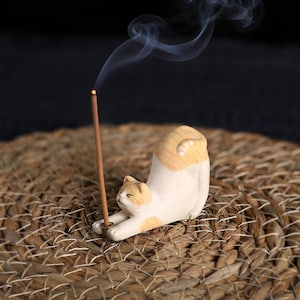 May include: A ceramic incense holder shaped like a stretching cat, with white and orange markings. The cat holds a lit incense stick, emitting a wisp of smoke. The holder rests on a woven surface, creating a calming and decorative item.