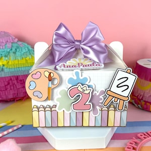May include: A white gift box decorated with art-themed stickers, a purple bow, and the name "Ana Paula." The box features a palette, paintbrush, number "2," and an easel with the number "3." The box is decorated with pastel-colored pencils.