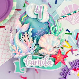Op de afbeelding: Een gelaagde taarttopper met een zeemeerminstaart, een parel in een open schelp en koraalrif-elementen. Het ontwerp bevat het cijfer 4 en een banner met de naam Camila in wit schrift. Het kleurenpalet bestaat uit pastelroze, blauwe, groenblauwe en paarse tinten met een felrood zeester-accent.