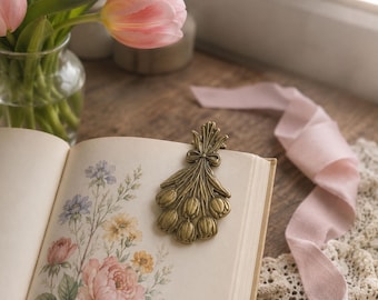 May include: A metallic gold-toned bookmark shaped like a bouquet of tulips with a small bow detail at the top. The bookmark is resting on the open page of a book that features a botanical illustration of various flowers in soft pastel colors.