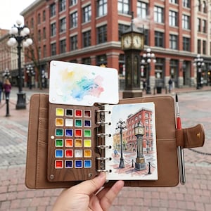 May include: A brown leather watercolor paint set with a wooden palette containing various colors. The set includes a small watercolor painting of a building with a clock tower and a watercolor brush. A white palette with watercolor paint is also included.