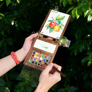 May include: A wooden watercolor palette with a built-in easel, holding a watercolor painting of fruit. The palette contains a variety of colorful paints and a small metal cup. A person is holding the palette and a paintbrush.
