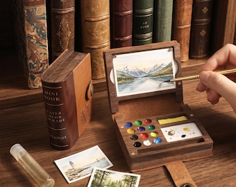 May include: A portable wooden watercolor set shaped like a book, featuring a built-in palette with twelve paint wells, a mixing area, and a small landscape painting. Includes a small water bottle and two additional miniature paintings.