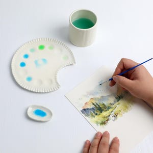 Op de afbeelding: Een aquarel in uitvoering, met een landschap met bergen en bomen. De kunstenaar gebruikt een blauwe penseel. Een wit keramisch palet met kleurrijke verf en een kopje turquoise vloeistof staan in de buurt.