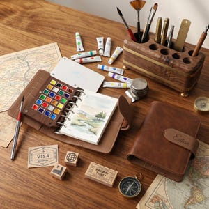 Könnte beinhalten: Eine Sammlung von Kunstbedarf, darunter ein braunes Lederjournal mit Aquarellfarben, Farbtuben und Pinseln. Ein kleines Notizbuch mit einer Landschaftsmalerei ist geöffnet. Weitere Artikel sind ein Kompass, Stempel und ein geschlossenes Lederjournal.