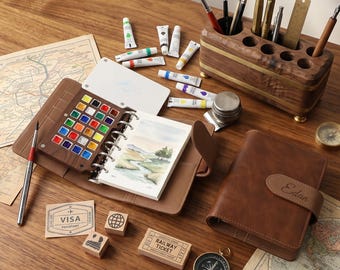 May include: A brown leather art journal with a watercolor palette, paint tubes, brushes, and a wooden pen holder. A small notebook with a landscape painting is open. Other items include a compass, stamps, and a closed leather journal.