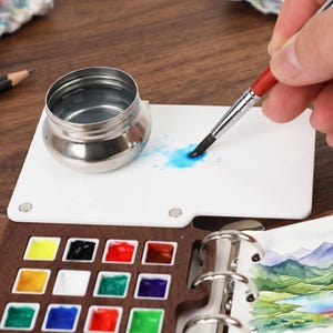 May include: A watercolor painting set with a wooden palette containing various paint colors. A small, silver water container and a white palette are also present. A paintbrush is applying blue paint to the palette. A pencil and a watercolor painting are also visible.