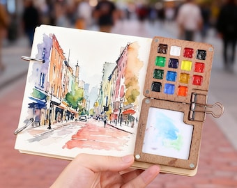 May include: A spiral-bound sketchbook featuring a watercolor painting of a city street scene. Attached to the side is a wooden palette with sixteen small square paint wells and a white mixing area, secured by a metal binder clip.