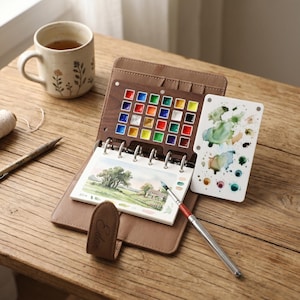 May include: A brown leather watercolor paint set with a wooden palette of colorful paints, a small watercolor pad with a landscape painting, and a white palette with paint splotches. A ceramic mug with floral designs sits nearby.