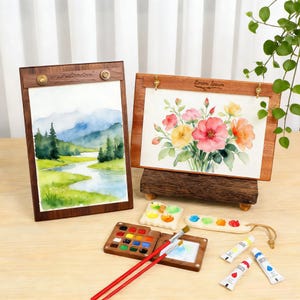 May include: Two watercolor paintings in wooden frames, one depicting a landscape and the other a floral arrangement. The scene includes miniature paint palettes, brushes, and paint tubes, suggesting an art supply theme.