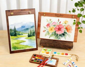 May include: Two watercolor paintings in wooden frames, one depicting a landscape and the other a floral arrangement. The scene includes miniature paint palettes, brushes, and paint tubes, suggesting an art supply theme.